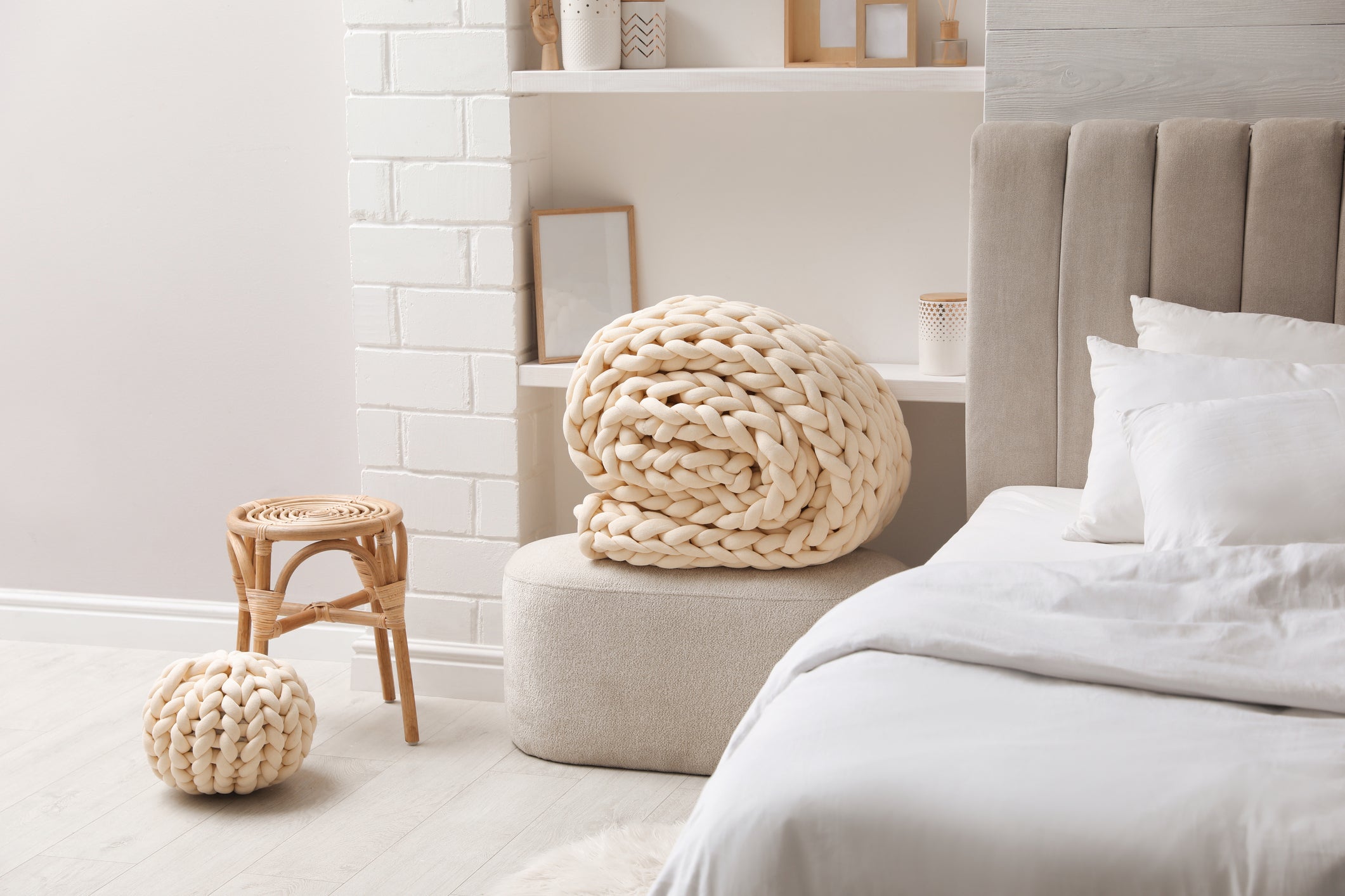Chunky knit weighted blanket in beige in a cozy minimalist bedroom with a taupe headboard, small stool, and natural decor.