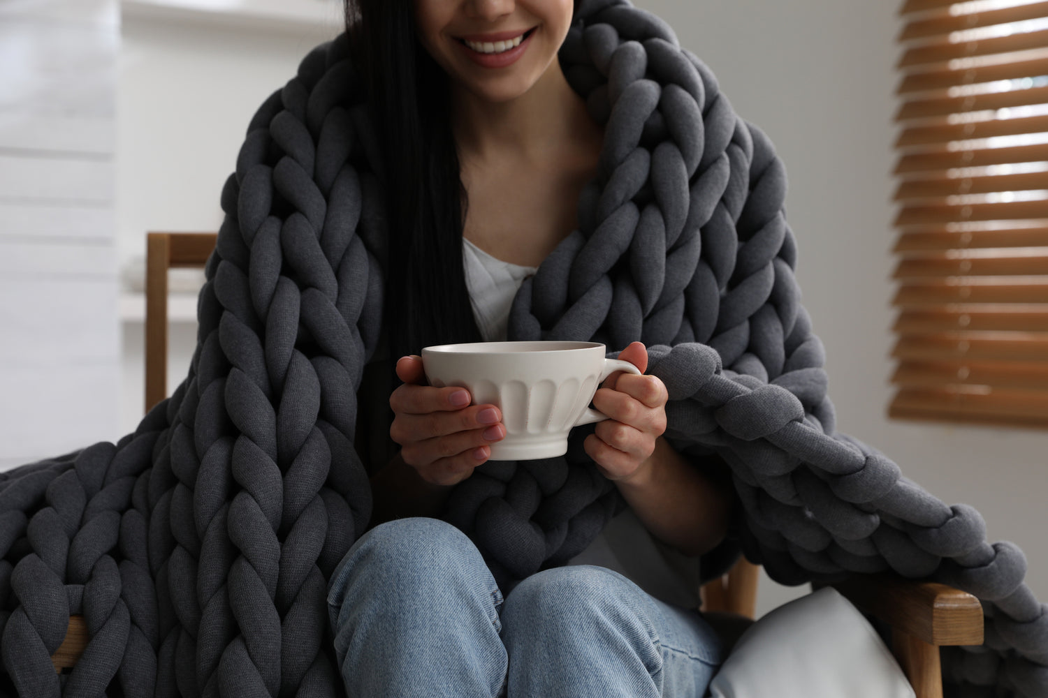 A woman wrapped in a chunky gray knit weighted blanket holding a cup of tea enjoying a calming moment. indoors