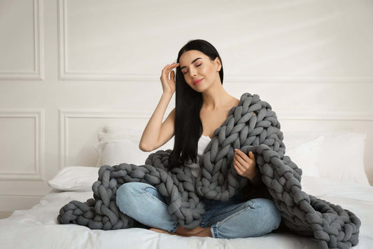 Woman sitting on a bed wrapped in a chunky gray cotton knit weighted blanket, looking relaxed and comfortable, demonstrating cooling, breathable comfort from Blanket & Bloom’s BloomKnit™ weighted blanket for menopause and hot flashes.