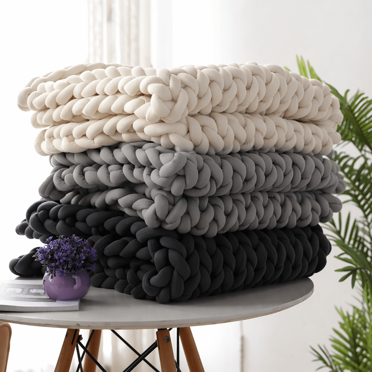 BloomKnit™ Chunky Cotton Weighted Blanket in various colors stacked on a table with a plant and purple vase.