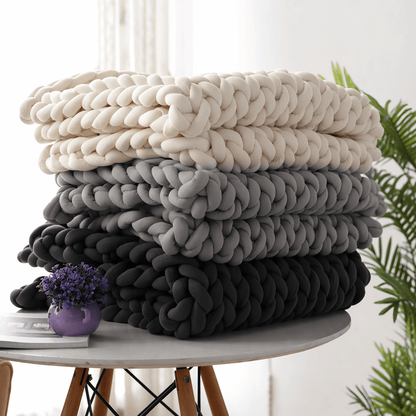 BloomKnit™ Chunky Cotton Weighted Blanket in various colors stacked on a table with a plant and purple vase.