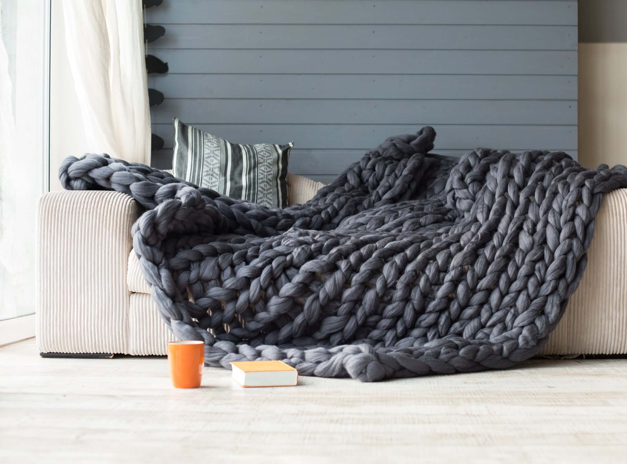 Gray chunky knit weighted blanket draped over a cream sofa in a cozy modern living room with natural light and warm tones.