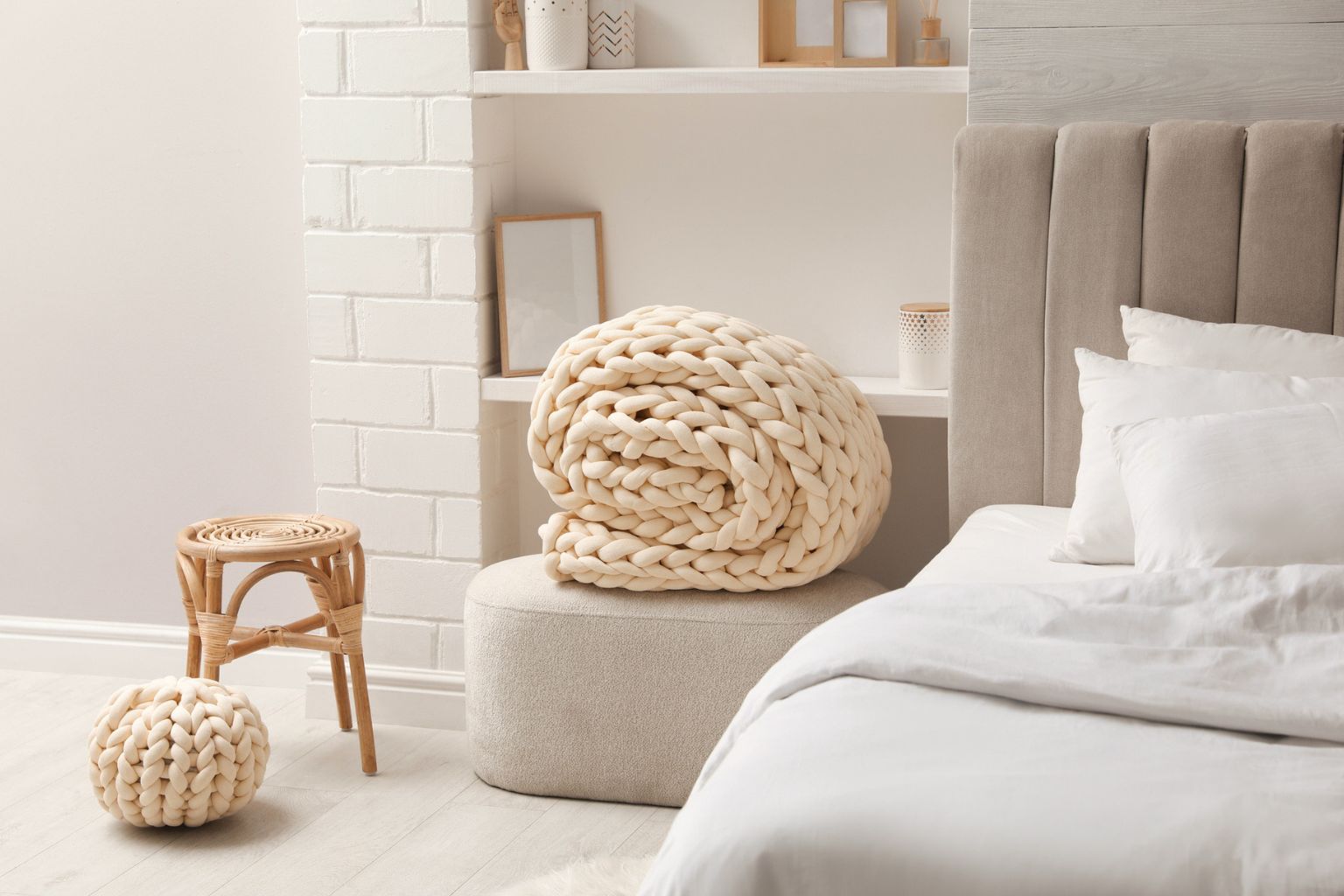 A chunky knit weighted blanket in a warm sand color in a bright bedroom setting next to a bed with white linens.