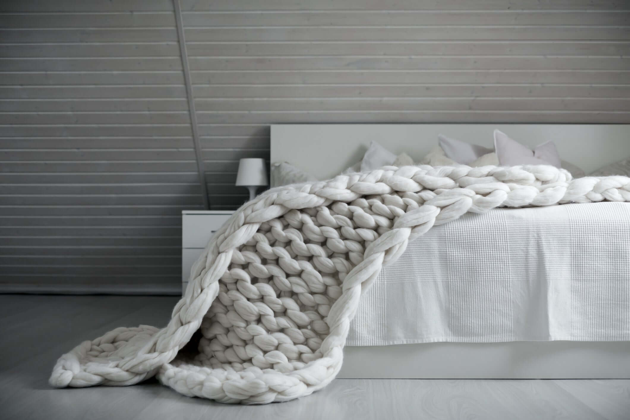 White chunky knit weighted blanket elegantly draped over a modern bed in a minimalist gray-toned bedroom.